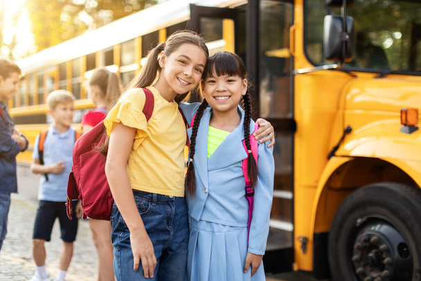 Boldog gyerekek pózolnak a sárga iskolabusz közelében, készen arra, hogy hazamenjenek az órák után, vidám, többnemzetiségű lányok hátizsákban ölelkezve és mosolyogva a kamerába, élvezve a barátságukat. - Fotó, kép