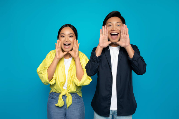 Happy asian couple in casual putting hands to mouth, screaming, isolated on blue studio background. People emotions from big sale, secret, gossip, attention and good news - Photo, Image