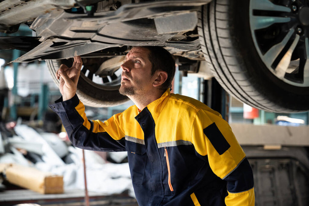 Araç tamircisi kaldırılmış aracın altından araba teftişi yapıyor. Üniformalı otomotiv teknisyeni araba akslarını ve şasi parçalarını dikkatle teşhis ediyor ve kontrol ediyor. Oxus. - Fotoğraf, Görsel