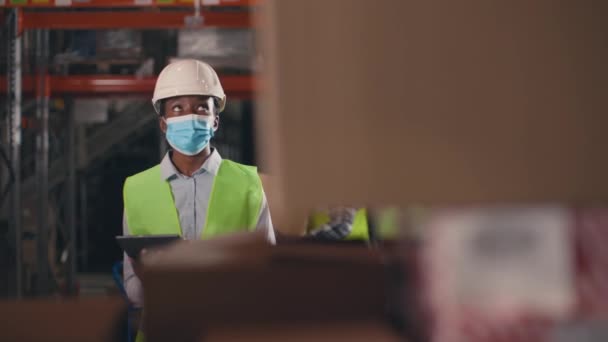 Afrikai amerikai nő a polcok mögött dobozokkal. Egy nő dolgozik a raktárban. Női tároló alkalmazott maszkban, védősisakban és sárga mellényben. Női tartó tabletta és az áruk kiszámítása. - Felvétel, videó