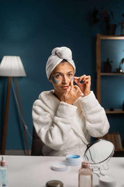 Millennial woman applying under eye patch on face and looking at camera. Beautiful woman wear bathrobe and wrapped bath towel on head. Face skin care. Copy space - Photo, Image