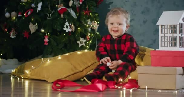 Infant with toy candle sitting amongst presents near Christmas tree in background. Child in Santa Claus hat waiting for Christmas to be spent at home - Footage, Video