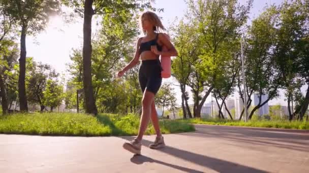schlanke Frau mit blonden lockigen Haaren in Aktivkleidung, die im morgendlichen Sonnenlicht zum Yoga-Training in die sommerliche Stadtlandschaft läuft - Filmmaterial, Video