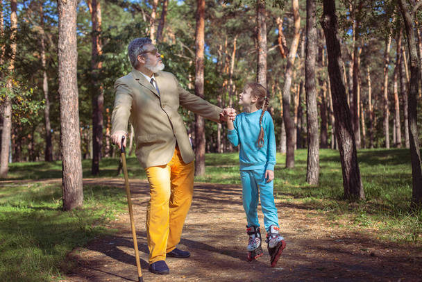 Grootvader loopt met zijn kleindochter in het park. Een meisje rolschaatst in het park met haar oude opa. Een grappige grootvader en een klein meisje spelen en maken grapjes.. - Foto, afbeelding
