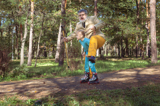 Grootvader loopt met zijn kleindochter in het park. Een meisje rolschaatst in het park met haar oude opa. Een grappige grootvader en een klein meisje spelen en maken grapjes.. - Foto, afbeelding