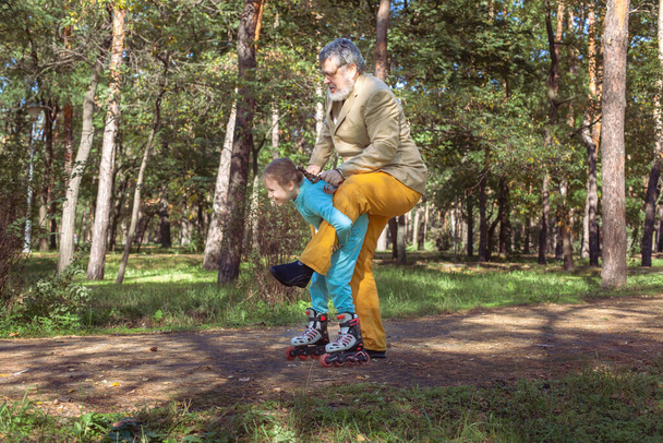 Nagyapa az unokájával sétál a parkban. Egy lány görkorcsolyázik a parkban az öreg nagyapjával. Egy vicces nagyapa és egy kislány játszanak és viccelődnek.. - Fotó, kép