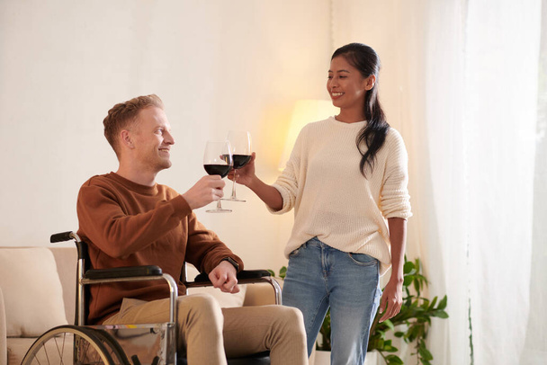 Jongeman in een rolstoel drinkt wijn met zijn vrouw thuis viert jubileum - Foto, afbeelding