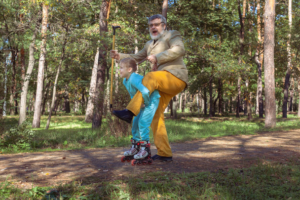 Grand-père marche avec sa petite-fille dans le parc. Une fille patine à roulettes dans le parc avec son vieux grand-père. Un grand-père drôle et une petite fille jouent et plaisantent. - Photo, image