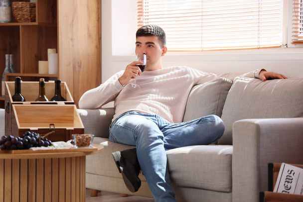 Νεαρός άνδρας με ποτήρι κόκκινο κρασί κάθεται στον καναπέ στο σπίτι - Φωτογραφία, εικόνα