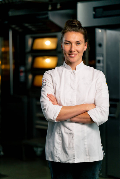 Portret van een lachende vrouwelijke chef-kok bakker in een professioneel uniform in een bakkerij tegen de achtergrond van een oven en vers gebakken goederen - Foto, afbeelding