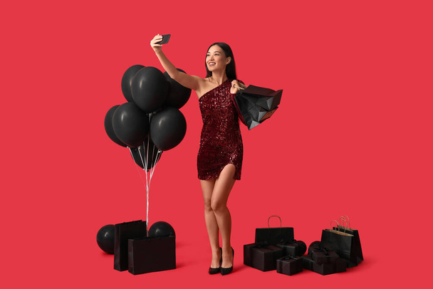 Krásná mladá asijská žena s dárkové krabice, nákupní tašky a balónky přičemž selfie na červeném pozadí. Černý pátek prodej - Fotografie, Obrázek