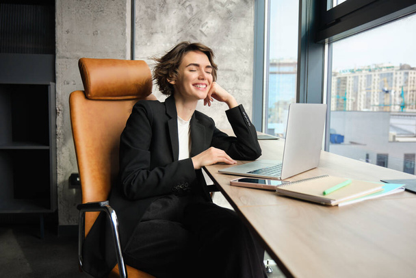Портрет бізнес-леді в офісі, менеджер з використанням ноутбука, робота над проектом, сидіння біля вікна в хмарочосі будівлі, очікування клієнта, в чорному костюмі. - Фото, зображення