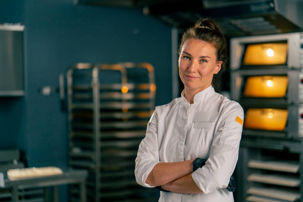 Een vrouwelijke bakker staat achter een professioneel keukenoppervlak in een bakkerij tegen de achtergrond van aangezette ovens en kijkt naar de camera - Foto, afbeelding