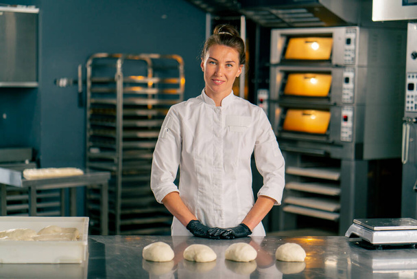 Femme boulanger se tient derrière une surface de cuisine professionnelle dans une boulangerie avec pâte préparée pour la cuisson et regarde la caméra - Photo, image