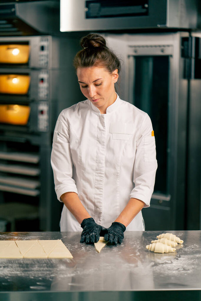 Ένα κορίτσι σεφ με σακάκι και γάντια σχηματίζει κρουασάν από ίσα μέρη ακατέργαστης ζύμης για πρωινό ψήσιμο σε φούρνο - Φωτογραφία, εικόνα