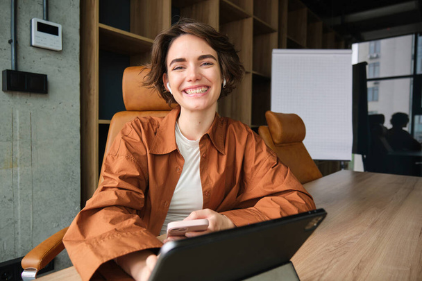Портрет молодой уверенной женщины на работе, начинающей предпринимательницы в повседневной одежде, сидящей в офисе с цифровым планшетом и улыбающейся в камеру. - Фото, изображение