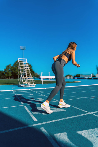 Krásná mladá opálená žena s dlouhými tmavými vlasy, připravena udělat sprint běží s nohama ohnutýma a v poloze na modré běžecké dráze. - Fotografie, Obrázek