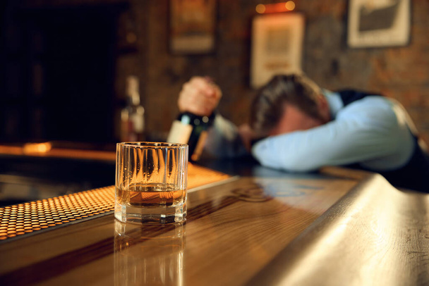 酒飲みの男がバーカウンターで寝て,ガラスでアルコール飲料を飲むことに集中する. ストレスのある日とアルコール依存症の概念 - 写真・画像