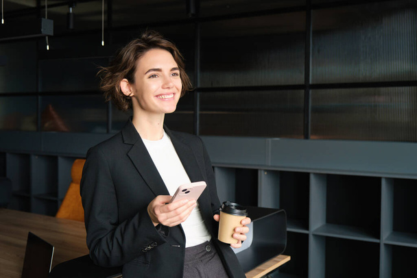 Ritratto di bella commessa in abito, in piedi con caffè e smartphone, sorridente, in posa in sala conferenze. - Foto, immagini