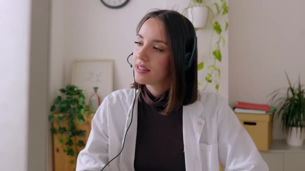 Portret młodej lekarki w słuchawkach, pracującej w pokoju konsultacyjnym, słuchającej pacjenta przez wirtualne połączenie wideo. Koncepcja pomocy medycznej. - Materiał filmowy, wideo