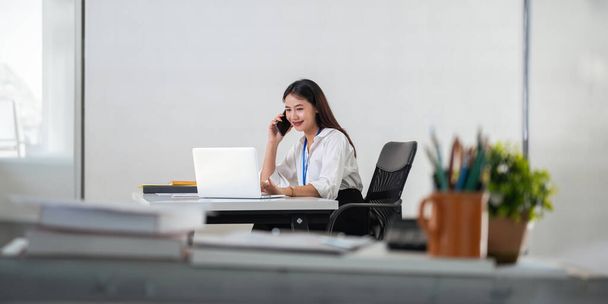 Šťastná mladá asijská podnikatelka mluvit na mobilním telefonu a usmívat se při sezení na pracovišti v kanceláři. - Fotografie, Obrázek