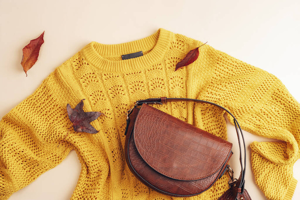 Vlakke lay van vrouwen gele trui en bruine zak, herfst bladeren op beige achtergrond. Herfst modeconcept. Bovenaanzicht, vlak lay. - Foto, afbeelding