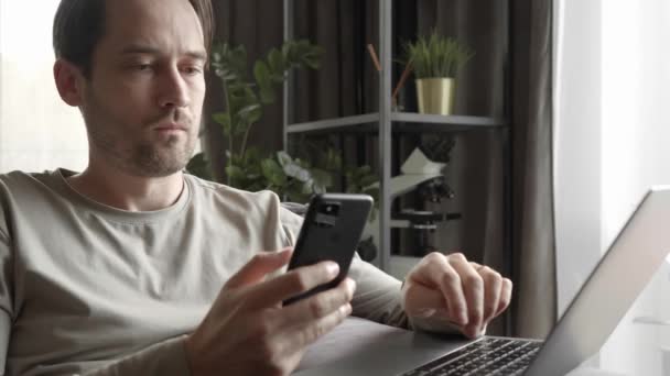 El hombre mira cuidadosamente a través y desplaza el contenido en la pantalla del teléfono inteligente, escribe texto en el teclado del ordenador portátil y utiliza el trackpad mientras está sentado en el sofá de la habitación. Trabajo remoto en casa. - Imágenes, Vídeo