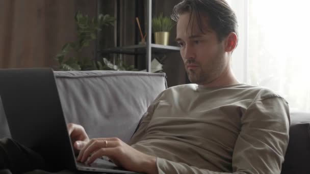 Un hombre está trabajando en un portátil, acostado en casa en el sofá. Un freelancer con ropa de casa mira fijamente a la pantalla del portátil y escribe texto en el teclado. Trabajo remoto y educación en línea en el hogar. - Imágenes, Vídeo