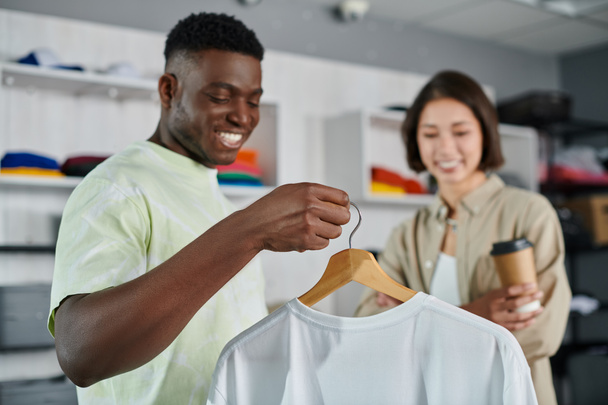 陽気なアフリカ系アメリカ人デザイナーが白いTシャツをプリントスタジオで笑顔のアジア人同僚に見せる - 写真・画像