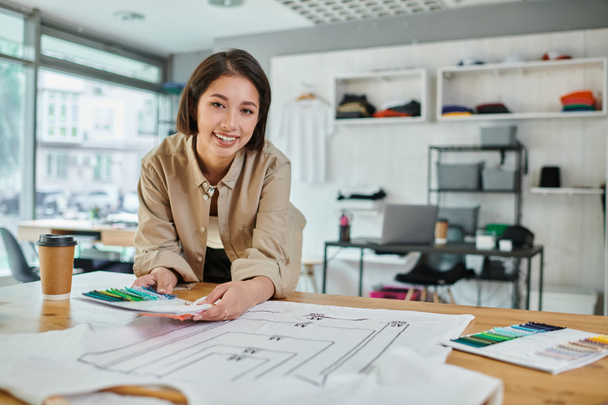 freudige kreative asiatische Designerin mit Farbmustern in der Nähe von Kleidung mit Formatgrößen im Druckstudio - Foto, Bild