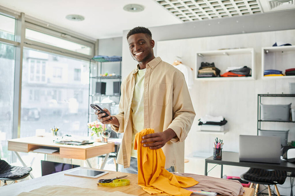 χαρούμενος αφρικανός Αμερικανός σχεδιαστής με smartphone και δείγματα υφάσματος κοιτάζοντας την κάμερα στο στούντιο εκτύπωσης - Φωτογραφία, εικόνα