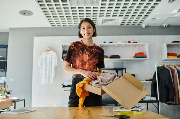веселий азіатський підприємець тримає картонну коробку з одягом і дивиться на камеру в друкованому ательє - Фото, зображення