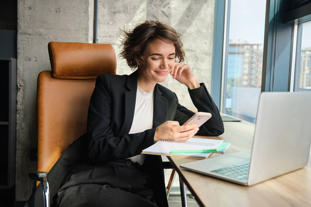 Retrato de mulher em um escritório, trabalhando em laptop, fazendo anotações, sentado perto da janela. - Foto, Imagem