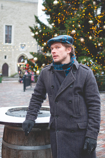 Ένας όμορφος νεαρός άνδρας με γκρι παλτό με σκωτσέζικο καπέλο τουίντ και ταρτάν φουλάρι που στέκεται σε μια πλατεία του Δημαρχείου του Ταλίν μια λαμπερή χειμωνιάτικη μέρα με ένα χριστουγεννιάτικο δέντρο και ένα χριστουγεννιάτικο εισιτήριο στο βάθος. - Φωτογραφία, εικόνα