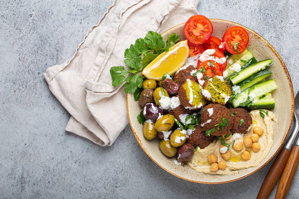 Falafel salad bowl with hummus, vegetables, olives, herbs and yogurt sauce. Vegan lunch plate top view on rustic stone background, healthy meal with falafel and veggies, space for text - Photo, Image