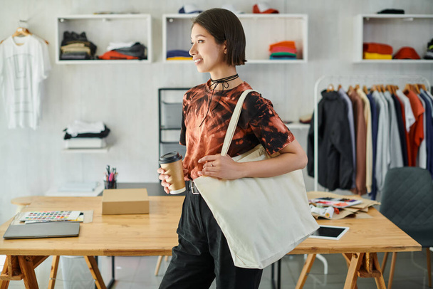 χαρούμενη και στιλάτη Ασιάτισσα με τσάντα για ψώνια και takeaway ποτό σε μοντέρνο στούντιο εκτύπωσης - Φωτογραφία, εικόνα