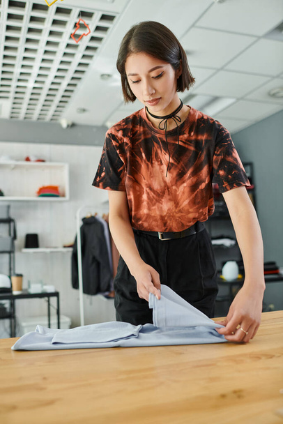 jovem e na moda asiático mulher dobrar roupas na mesa em têxtil imprimir estúdio, pequeno negócio - Foto, Imagem