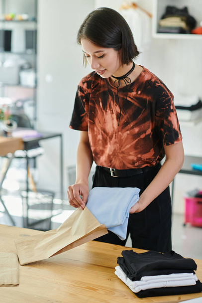 jong en stijlvol aziatisch vrouw verpakking gevouwen kleding in papieren zak in print studio, klein bedrijf - Foto, afbeelding