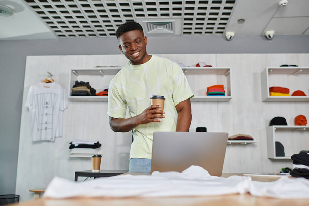χαμογελαστός Αφροαμερικάνος σχεδιαστής με καφέ για να πάει κοιτάζοντας το φορητό υπολογιστή κοντά στα ρούχα στο στούντιο εκτύπωσης - Φωτογραφία, εικόνα