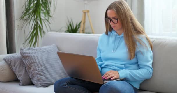 Nuori nainen istuu sohvalla kannettavan tietokoneen avulla katsoen näytön kirjoitusviestiä, nainen chattailee tietokoneella, selailee Internetin sosiaalista mediaa opiskelee tai työskentelee verkossa kotona - Materiaali, video