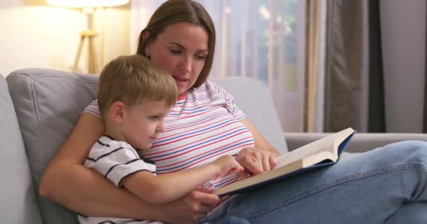 Matka a syn si čtou knížku, jak sedí na útulné pohovce v obýváku, v rodinném čase. Vysoce kvalitní 4K záběry - Záběry, video