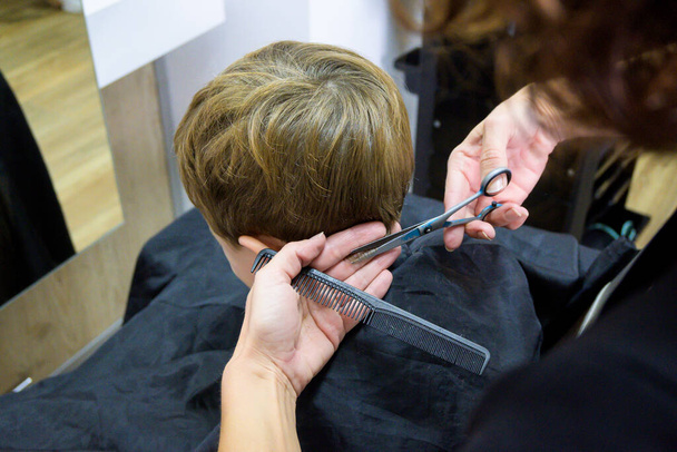 Um menino bonito senta-se em um cabeleireiro nos estilistas, uma criança em idade escolar está recebendo corte de cabelo em um salão de beleza, uma criança em uma barbearia, um corte de cabelo homens curtos. - Foto, Imagem