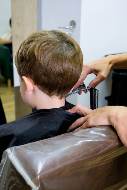 Um menino bonito senta-se em um cabeleireiro nos estilistas, uma criança em idade escolar está recebendo corte de cabelo em um salão de beleza, uma criança em uma barbearia, um corte de cabelo homens curtos. - Foto, Imagem