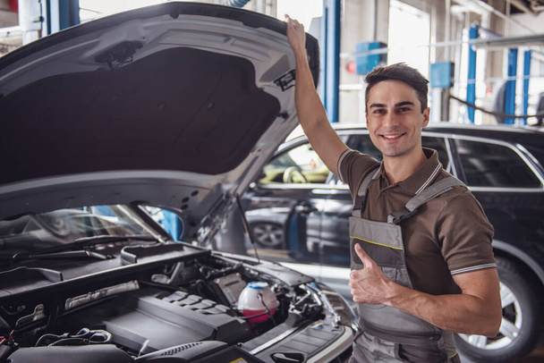 Handsome young auto mechanic in uniform is showing Ok sign, looking at camera and smiling while examining car in auto service - Photo, Image