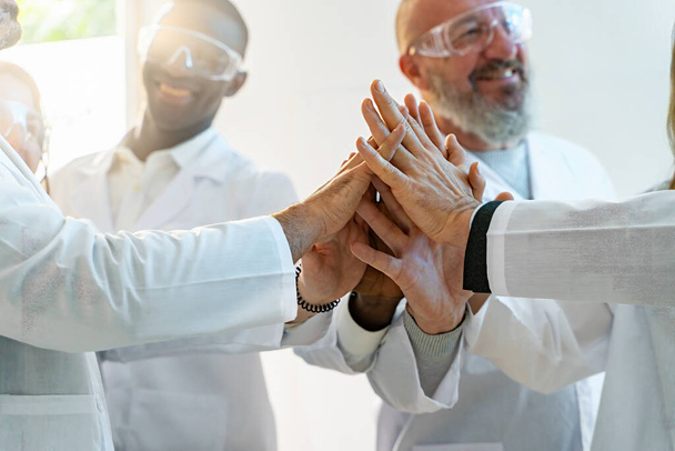 Een groep chemici, met een beschermende bril op, bundelen hun handen in eenheid. De focus ligt sterk op hun handen en symboliseert teamwork en betrokkenheid op wetenschappelijk gebied.. - Foto, afbeelding
