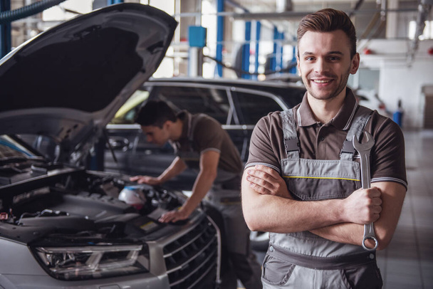 At the auto service. Handsome young auto mechanic in uniform is looking at camera and smiling while his colleague is examining car - Photo, Image