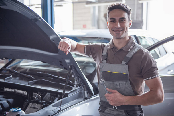 Atractivo joven mecánico de automóviles en uniforme está mostrando signo Ok, mirando a la cámara y sonriendo mientras está de pie en el servicio automático
 - Foto, imagen