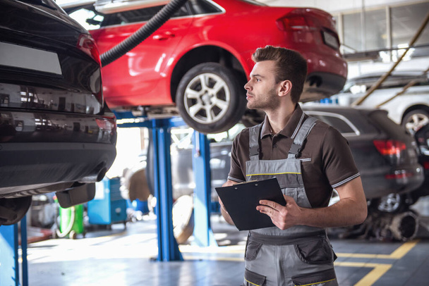 Handsome young auto mechanic in uniform is making notes while examining car in auto service - Photo, Image