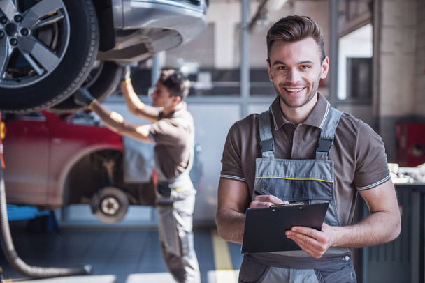 At the auto service. Handsome young auto mechanic in uniform is holding a folder, looking at camera and smiling while his colleague is examining car - Photo, Image