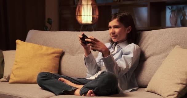 Der kleine Junge sitzt auf dem Sofa, hält ein Gamepad in den Händen und ist tief in die virtuelle Welt der Videospiele vertieft. Seine Augen sind auf den Bildschirm gerichtet, Finger manövrieren gekonnt die Bedienelemente.. - Filmmaterial, Video